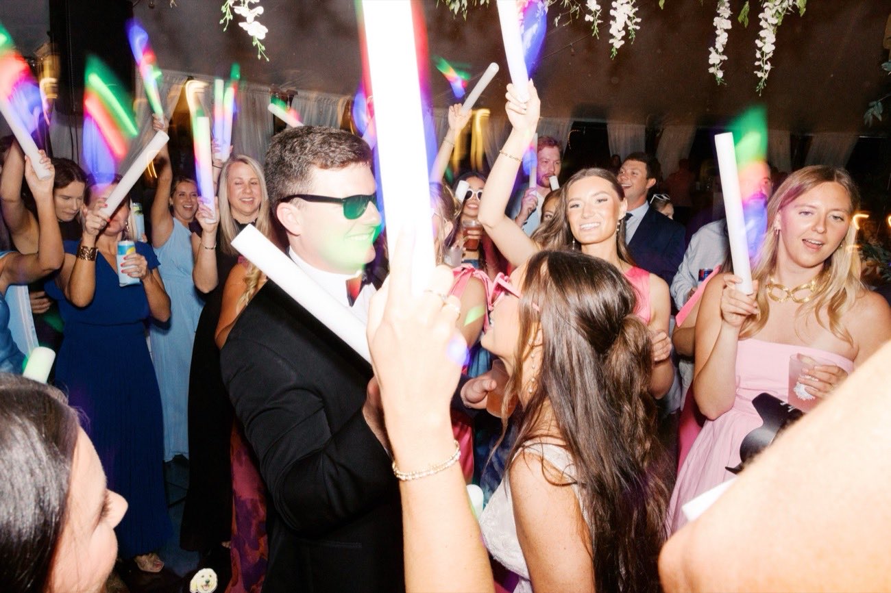Groom celebrating with guests holding oversized glow sticks under a floral arch